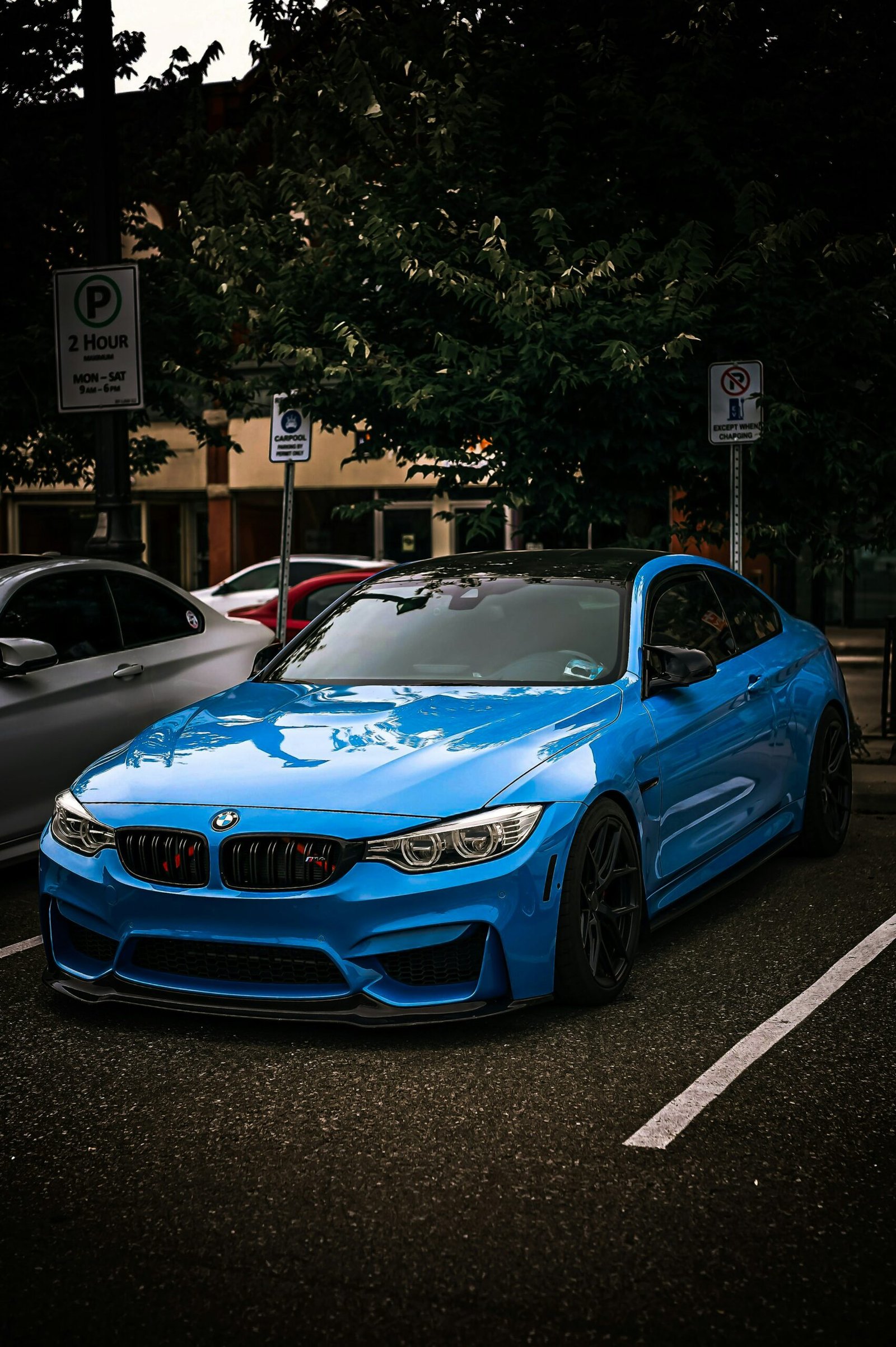A bright blue BMW M4, with a black roof and black wheels, is parked in a paved parking lot with white lines. Its red and black grille stands out. Behind it, lush green trees provide a dark backdrop, partially obscuring other parked cars. To the left, a 