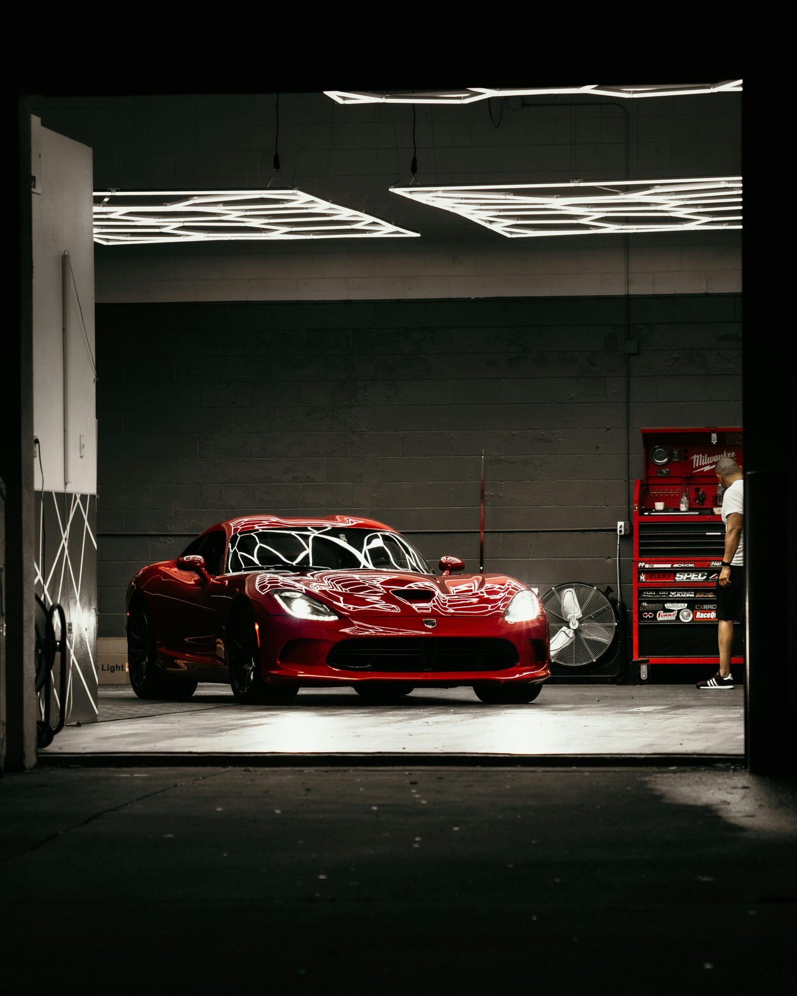 A sleek, glossy red Dodge Viper is prominently displayed in a dimly lit garage or detailing bay, its highly reflective surface mirroring the bright, rectangular overhead LED lights. The car is positioned in the center, facing the viewer. To the right, a person in a white t-shirt stands near a red tool chest and a large fan. The overall atmosphere is dark and dramatic, with the car serving as the luminous focal point.