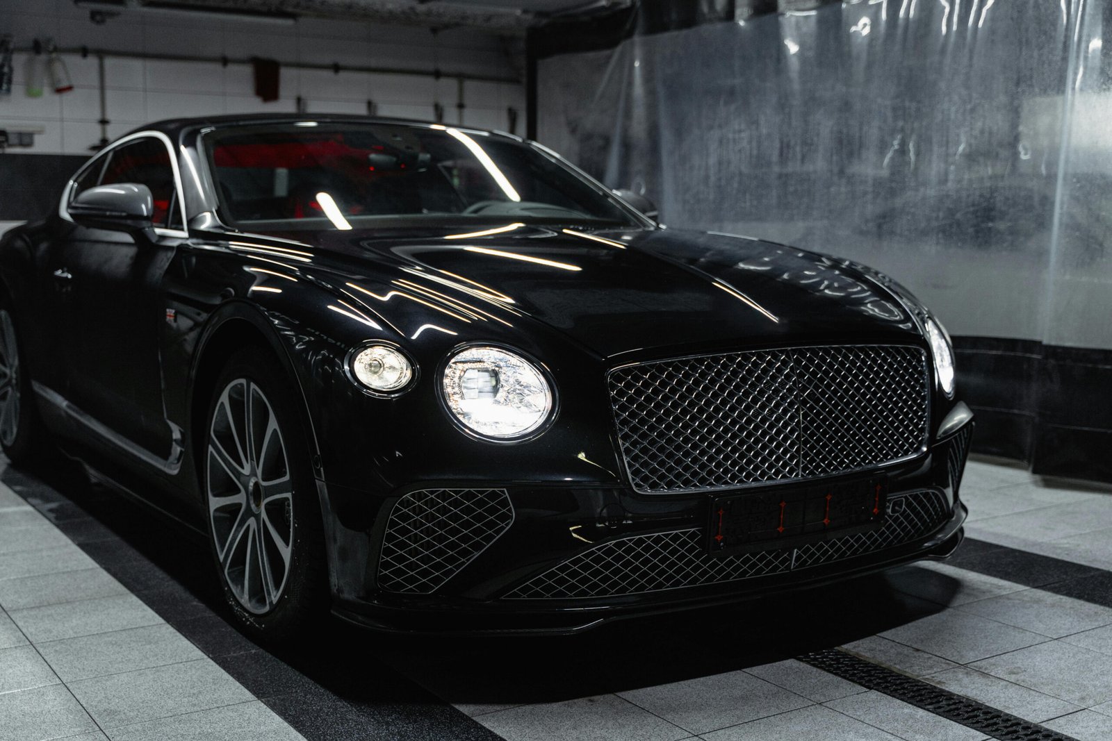 A luxurious black Bentley Continental GT is parked indoors, likely in a detailing bay or garage. Its highly reflective paint surface reflects the surrounding environment. The front of the car, with its distinctive large grille and circular headlights, is prominently featured. There are grey and black striped floor tiles beneath the car, and a clear or translucent plastic sheet hangs in the background.