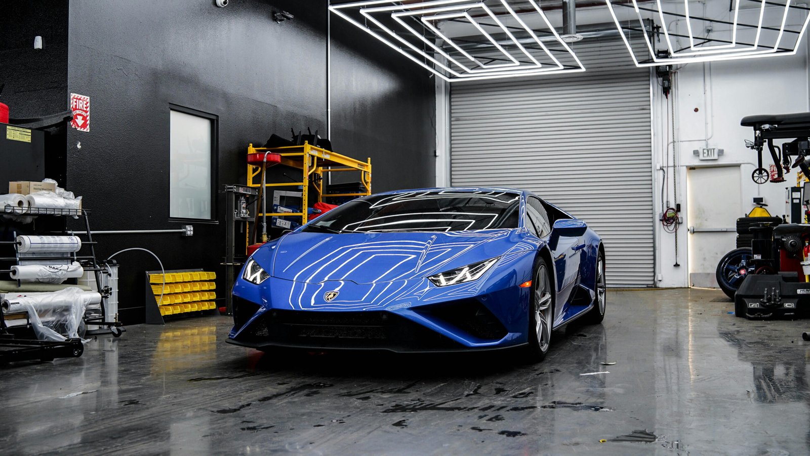 A vibrant blue Lamborghini Huracán parked in a modern garage or detailing shop. The car is centrally positioned, reflecting the geometric overhead LED lights on its glossy hood and roof. The background features black walls, a grey roll-up door, a window, yellow shelving with various items, and automotive equipment on the right side.
